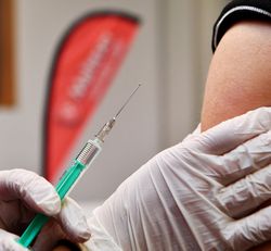 Das Foto zeigt einen Impfvorgang in Nahaufnahme. D.h. einen Oberarm, der von einer behandschuhten Hand gehalten wird sowie daneben eine aufgezogene Spritze in der anderen behandschuhten Hand des Impfenden. 